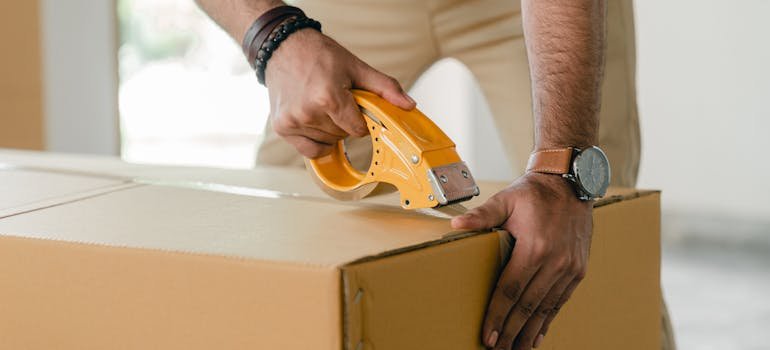 Tips For Moving A Grand Piano In Cincinnati 2 A person taping a cardboard box after filling it while moving a grand piano.
