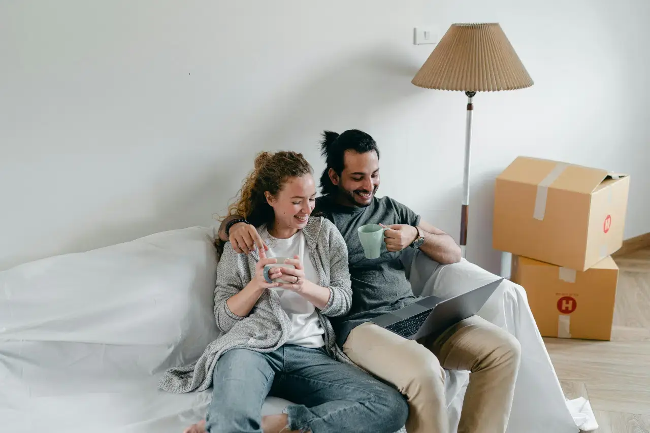 Happy couple sitting on a couch with coffee mugs, relaxing after unpacking in their new home with moving boxes in the background.