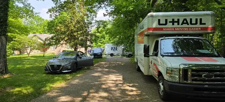 Commercial Movers 3 A U-Haul truck parks alongside a car on a tree-lined driveway leading to a house, indicating a moving day scenario.