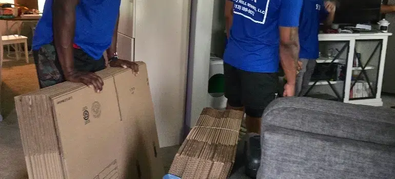 Three people wearing blue shirts stand indoors, holding large flat cardboard boxes and bundles of flattened moving boxes near a gray couch.