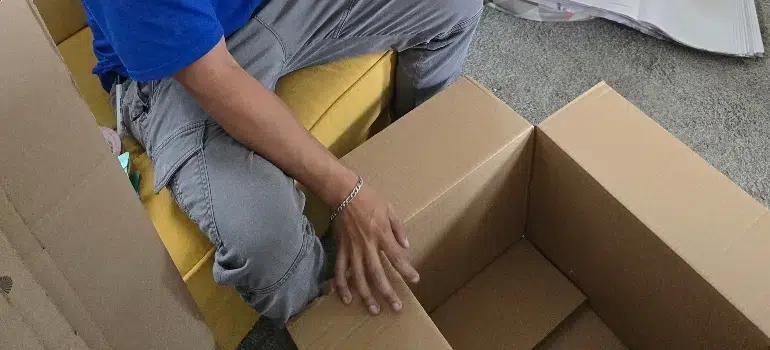 A person in a blue shirt and gray pants sits on a yellow sofa, assembling a cardboard box on a gray carpet. The scene conveys focus and productivity.