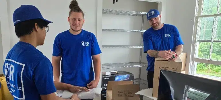 Three individuals in blue shirts are working in a bright room, packing items into cardboard boxes while discussing tasks.
