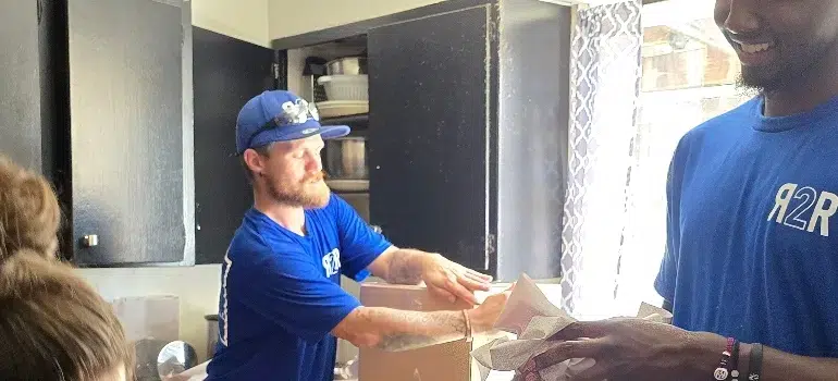 Two individuals in blue shirts unpack a box in a kitchen, surrounded by household items and sunlight streaming through a window.