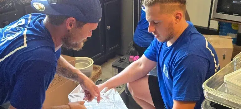 Two individuals in blue shirts are engaged in an activity involving a reflective surface, surrounded by cardboard boxes and containers.