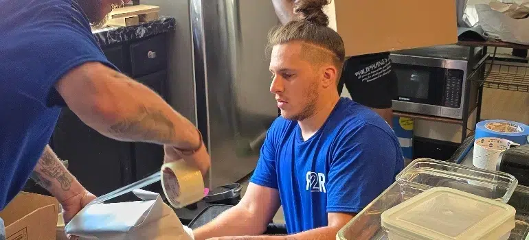 Two individuals in a kitchen are packing items into boxes, one using tape while the other sits nearby, surrounded by containers and a microwave.