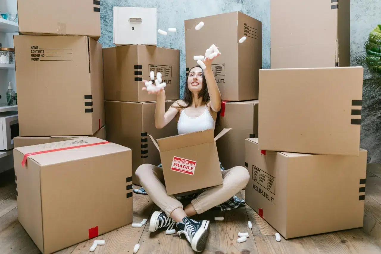 Woman sitting on the floor tossing packing peanuts, surrounded by stacked moving boxes.