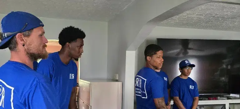 Fairfield Movers 5 A group of four men in blue shirts stands indoors, engaged in conversation next to a large TV and a white cabinet.