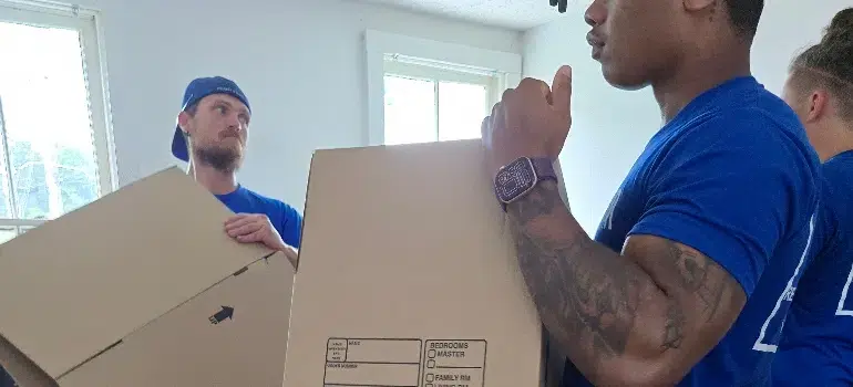 Two men in blue shirts carry moving boxes in a well-lit room with white walls and a window. The mood appears focused and collaborative.