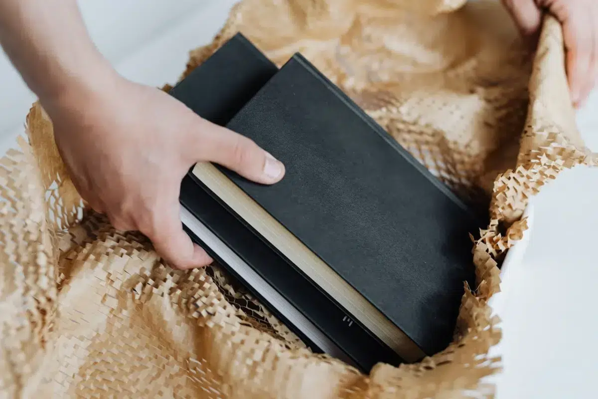 A hand carefully lifts black-bound books from a box filled with crumpled packing paper, showing the best way to pack books for moving