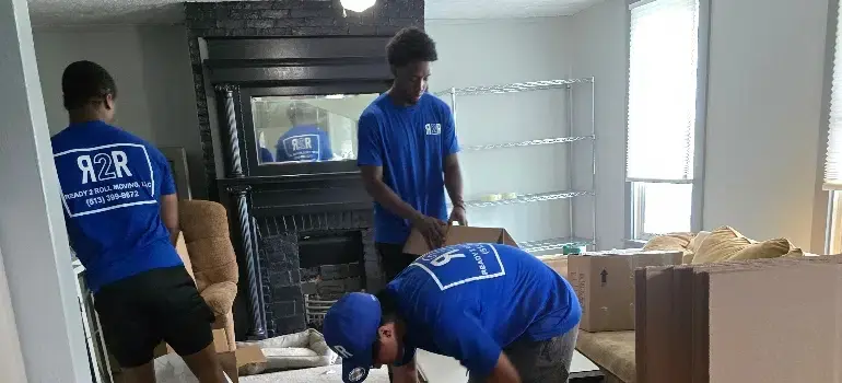 Three movers in blue shirts are packing boxes in a bright living room with a fireplace and shelves. A couch is visible nearby.