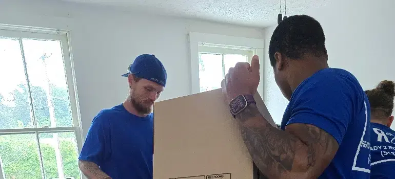 Residential Movers 5 Two movers in blue shirts are working together to lift a cardboard box inside a bright room with large windows.
